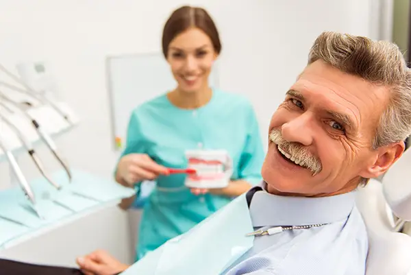 Smiling older patient in dental chair as hygienist demonstrates how he should brush his dentures 